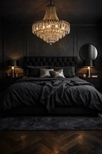 A moody black bedroom with a black upholstered bed, black bedding, a crystal chandelier overhead, and black nightstands with brass lamps.
